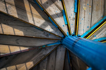 High angle view of an old spiral staircase with wooden steps and a bright blue metal central pole illuminated by sunlight creating a geometric abstract texture background with deep shadows