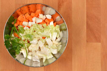 An overhead shot shows a metal bowl filled with diced carrots, chopped cabbage, cauliflower pieces, and fresh parsley, resting on a wooden surfac