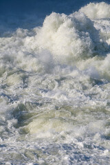 Strong powerful stream of rushing river water flows fast creating white foam and splashes against the deep blue background for a beautiful abstract natural texture of liquid energy outdoors