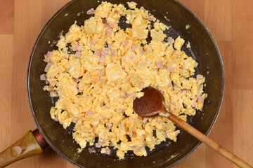 An overhead shot shows a dark frying pan containing fluffy scrambled eggs mixed with small pieces of ham and cheese, being stirred with a wooden spoon on a wooden background