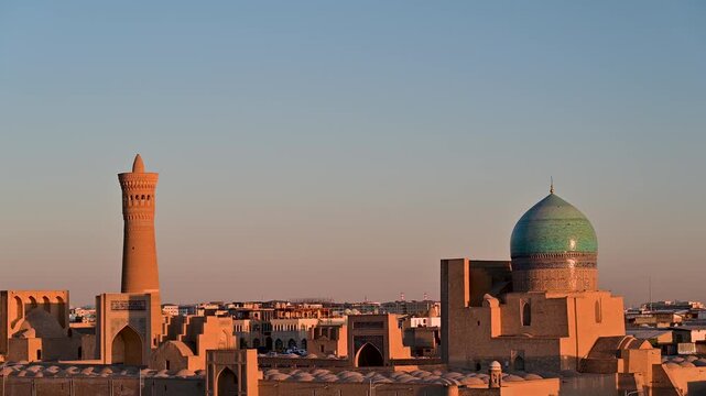 Warm golden hour light falls across the rooftops and historic structures of Bukhara, Uzbekistan, revealing the colors and textures of the old city in soft evening tones.