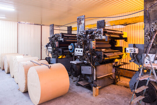 Large rolls of paper stand on the floor near heavy yellow industrial printing press machines inside a factory workshop illustrating the automated polygraphy production process workflow