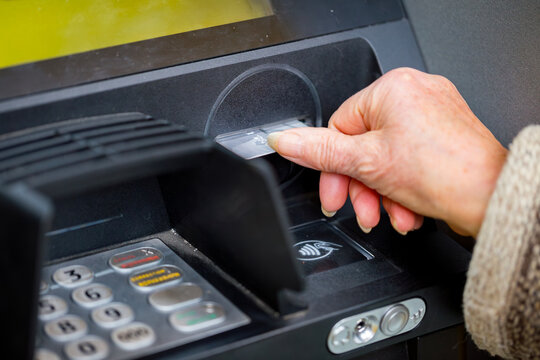 Close up view of an elderly woman hand correctly inserting a credit card into the black ATM machine reader slot to withdraw cash money from the bank account balance successfully outdoors