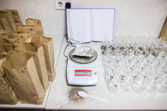 Electronic analytical balance scale stands on the white table near brown paper bags and glass flasks with soil samples ready for chemical agrochemical analysis research in the laboratory