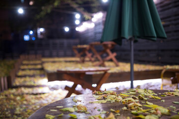 Empty wooden tables covered with yellow dry leaves stand on the outdoor cafe terrace at night under the blurred garland lights creating a cozy melancholic autumn atmosphere of the season end