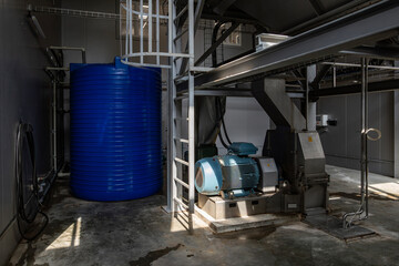 The interior of an industrial plant room. A large blue electric motor powers machinery next to a large blue plastic vertical storage tank. A metal ladder provides access to the equipment.