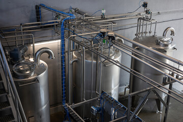 The interior of a modern industrial plant, such as a food, beverage, or chemical factory. Large stainless steel tanks are connected by a complex system of pipes, valves, and sensors.