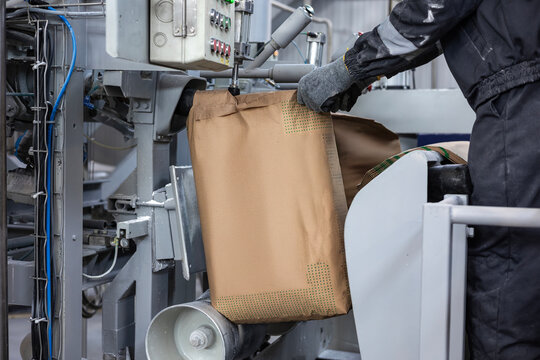 A factory worker in gloves operates an automated bagging machine. The industrial equipment is filling a large brown paper sack with bulk material. The worker guides the bag on the production line.