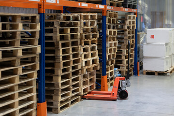 An orange manual pallet jack is parked on a concrete floor next to stacks of empty wooden pallets. The pallets are stored under tall blue and orange industrial racks in a modern warehouse.