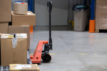 Fototapeta premium An orange manual pallet jack is parked on a concrete warehouse floor. This logistics equipment is next to a stack of cardboard boxes and a plastic pallet, ready for shipping.