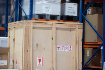 A large wooden shipping crate with fragile and keep dry logistics symbols sits in a modern warehouse. The background shows tall, blue and orange industrial racks filled with cardboard boxes on pallets © Sodel Vladyslav