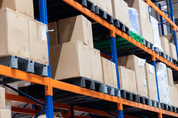 Cardboard boxes are stacked on tall, orange and blue industrial racking in a modern warehouse. The boxes, marked with handwritten numbers, are stored on plastic pallets.