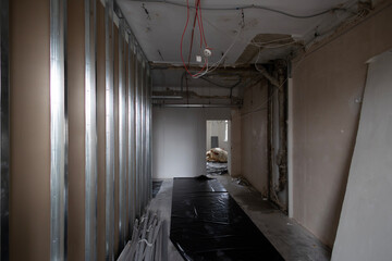 A under construction during an office renovation. New drywall partitions and metal studs form the walls. The floor is covered in black plastic. The open ceiling reveals exposed wires and cable trays.