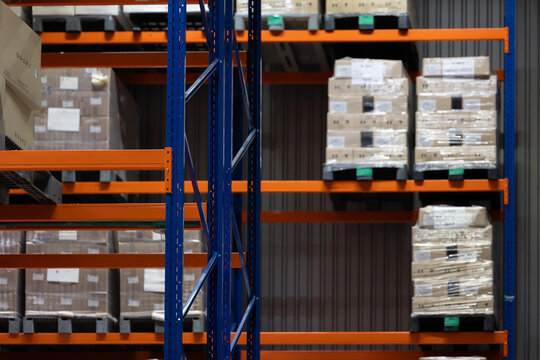 A modern warehouse interior with tall, blue and orange industrial racking. The shelves are filled with pallets, which hold cardboard boxes wrapped in plastic film, ready for shipping.