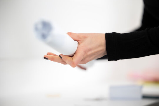 A woman's hand, with black nail polish, holds a rolled-up white paper document. This could be an architectural blueprint, a technical drawing, or a plan being reviewed in a bright office.