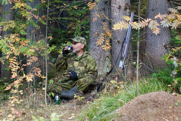 A hunter drinks tea under a tree in the forest