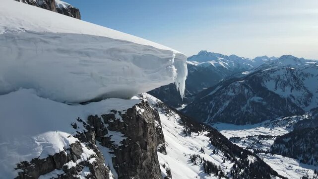 Snow Cornice Over Cliff - A delicate snow cornice overhanging a sharp cliff edge. The video showcases a mountain range and valley in the distance, covered in snow under a clear blue sky.
