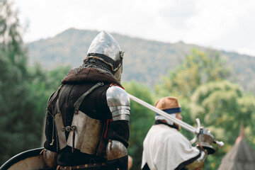 An ancient knight in armor holds a shield in his hand, and his armorer holds a great sword.