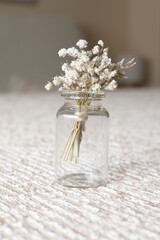 Small bouquet of delicate dried white flowers tied with twine in a clear apothecary jar, minimalist rustic still life with warm, muted tones and soft textured background