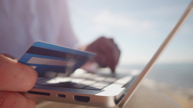 A digital nomad holds a credit card near a laptop, symbolizing remote work and financial transactions. The serene, possibly coastal setting emphasizes the freedom and flexibility of freelance work.