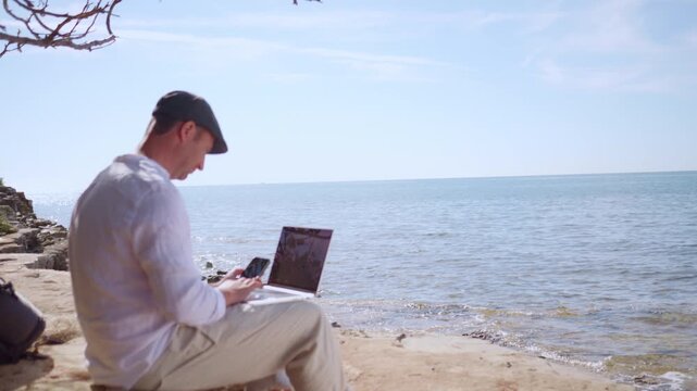 A focused digital nomad works on his laptop by the serene sea, embodying remote work freedom. The clear sky and tranquil waters highlight the peaceful, portable work environment.