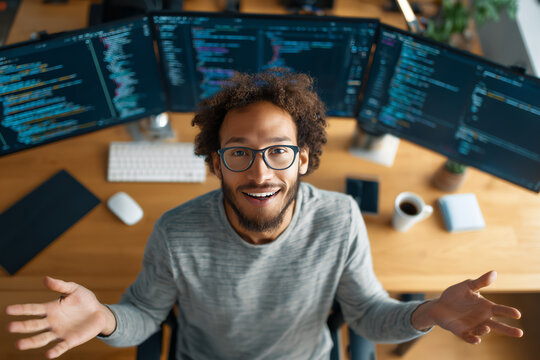 Happy young software developer smiling at a multi-monitor workstation while coding from a cozy home office with coffee and modern desk setup