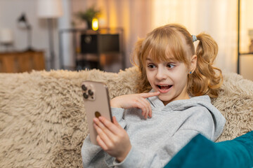 Young teenager girl sits at home scrolling pictures on a smartphone, laughing at funny memories. Blonde child kid on sofa points at the screen, chatting about family adventures with big smiles.