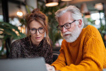 Multigenerational colleagues collaborating on a laptop in a cozy café — senior mentor guiding a younger professional through planning and problem-solving
