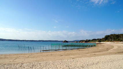 穏やかな青空の下、広がる砂浜と浅瀬の海面に、海苔養殖用のネットが並ぶ三重県の次郎六郎海岸