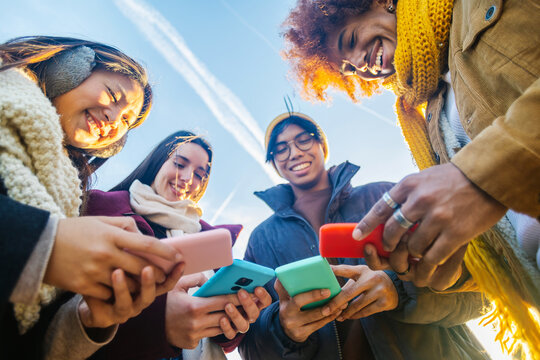 Four cheerful young students are using their smartphones together on a sunny winter day, enjoying online communication and social media