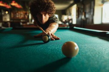 Young man lining up a precise pool shot on a green felt billiards table in a dimly lit bar — a focused moment of concentration, skill, and leisure