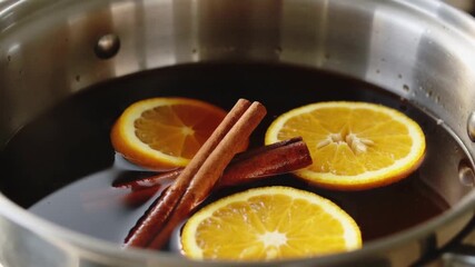 The video is a close-up of a pot filled with dark liquid mulled wine or a similar hot drink. Orange slices and cinnamon sticks float in the liquid. - Powered by Adobe