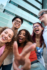 Vertical. Five cheerful friends take a playful selfie in a modern city, laughing and flashing peace signs by glass towers. Bright daylight, casual fashion, carefree mood, and vibrant diversity.