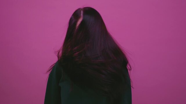 Beautiful cheerful brunette young girl wearing green pink sweater posing isolated on pink wall. Dancing shaking head with flowing hair. Teenage woman shakes her straight black long hair, slow motion.