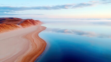 An aerial view of a tranquil coastline at sunrise, featuring rolling sand dunes meeting a calm, reflective ocean shrouded in mist.