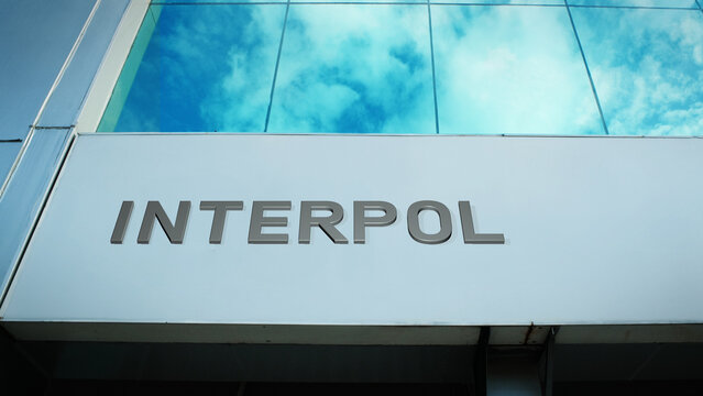 Interpol sign displayed on a modern glass building with windows reflecting clouds and sky, creating a bright, sleek, contemporary exterior with clear visual impact