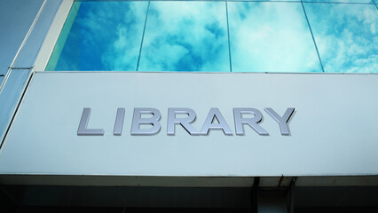 Library sign displayed on a modern glass building with windows reflecting clouds and sky, creating a clean, bright, contemporary exterior with an educational atmosphere