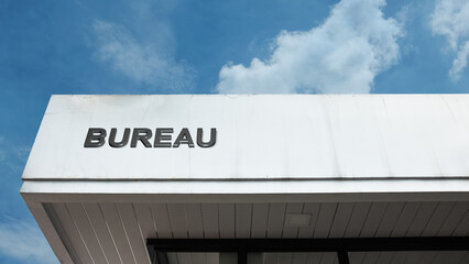 A bureau word sign mounted on a building facade under a clear blue sky, showing clean lettering, bright daylight, and a simple architectural exterior