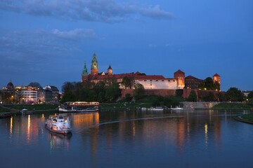 Historic Wawel Castle complex and Vistula River with blurred cruise ship in foreground in night illuminations. Krakow, Poland