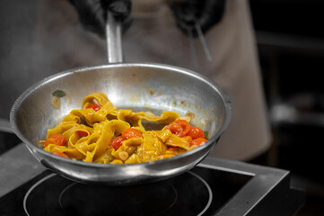 A chef wearing black gloves is stir-frying fresh, wide yellow pasta with cherry tomatoes and a vibrant sauce in a stainless steel pan on an induction hob.
