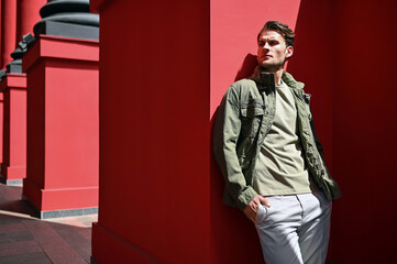 A man in a green jacket and light pants poses outdoors near a bright red wall. The confident stance and clean background highlight a modern casual style.