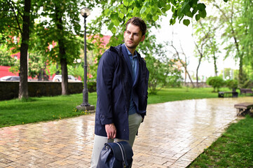 A young man in a navy coat walks through a park after the rain. Soft natural light, fresh greenery, and a relaxed pose create a calm and stylish urban mood.