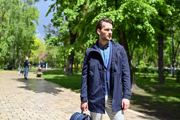 A stylish man walks through a sunlit park wearing a navy jacket and light trousers. Fresh greenery, soft shadows and his relaxed stride create a calm lifestyle atmosphere.