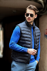 A fashionable man adjusts his sunglasses while wearing a gray padded vest over a bright blue shirt. The close-up shot highlights confidence and clean contemporary menswear style.