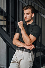 A man in a green T-shirt poses against a concrete wall, standing casually with one knee bent. The raw texture of the background adds a bold, contemporary fashion edge.