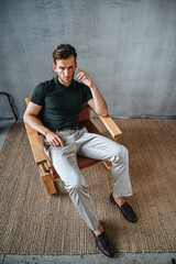 A man in a green T-shirt poses against a concrete wall, standing casually with one knee bent. The raw texture of the background adds a bold, contemporary fashion edge.