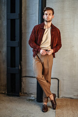 A full-body portrait of a man in a rust-colored jacket and beige trousers, standing in dramatic directional light. The mix of soft shadows and clean styling enhances the editorial feel.