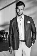 A stylish portrait of a man in a classic light suit, standing confidently in a softly lit interior. The monochrome look highlights texture, structure, and elegant formal fashion appeal.