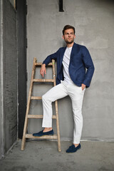 A man in a navy blazer stands by a wooden ladder in a textured concrete room, blending modern fashion with rustic industrial style.