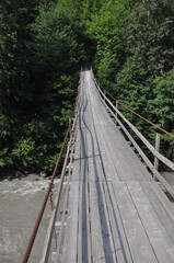 Obraz premium Wooden suspension bridge stretches over a river surrounded by lush greenery in summer